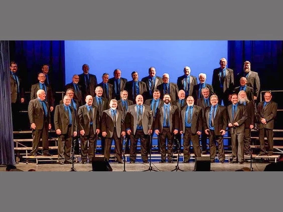 A men's choir stands on risers, dressed in formal black suits with blue ties, posing in a concert hall setting. The group is likely preparing for or has just finished a performance.