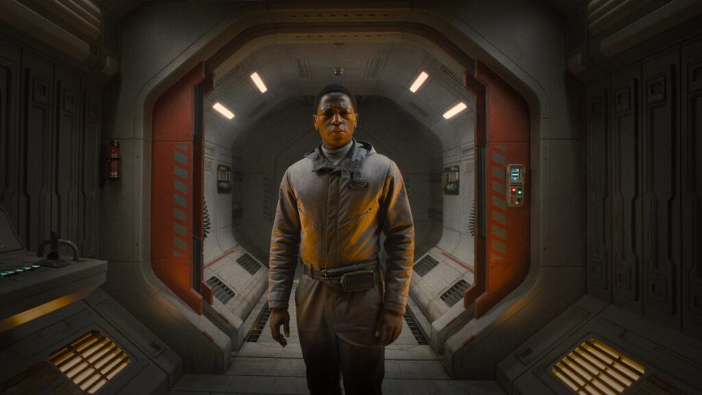 A scene from Alien: Romulus featuring actor David Jonsson. He stands in the middle of a futuristic, dimly lit spacecraft corridor wearing a grey spacesuit. The corridor is sleek, with metallic walls, red doors, and panels with control consoles. Jonsson's character appears serious and focused as he looks straight ahead.