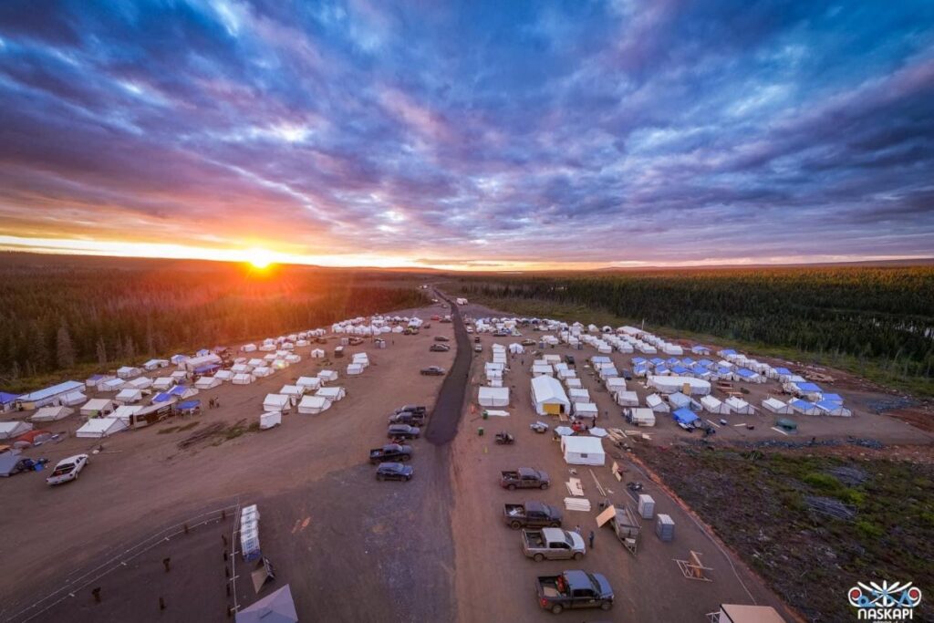 An aerial view of a large campsite at sunset shows rows of white tents spread across a vast open area. Trucks are parked near the tents, and a central road divides the camp. The sun is setting in the distance, casting a warm orange and yellow glow over the forest that surrounds the camp. The sky is filled with dramatic clouds, creating a peaceful yet majestic scene. The logo in the bottom right corner indicates the event is associated with the Naskapi.