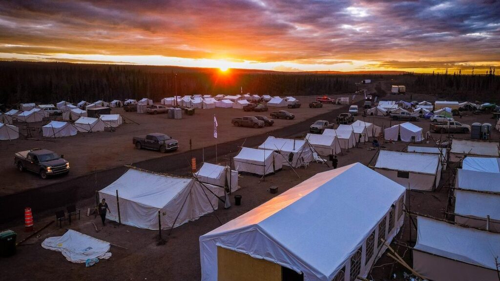 A campsite with multiple white tents is spread out over a flat area, surrounded by a forest of evergreen trees. The sunset paints the sky in vibrant hues of orange and yellow, casting a warm glow over the camp. Trucks are parked around the tents, and a few people are walking in the camp, preparing for the evening. The scene captures the peacefulness of the camp as the day comes to a close.