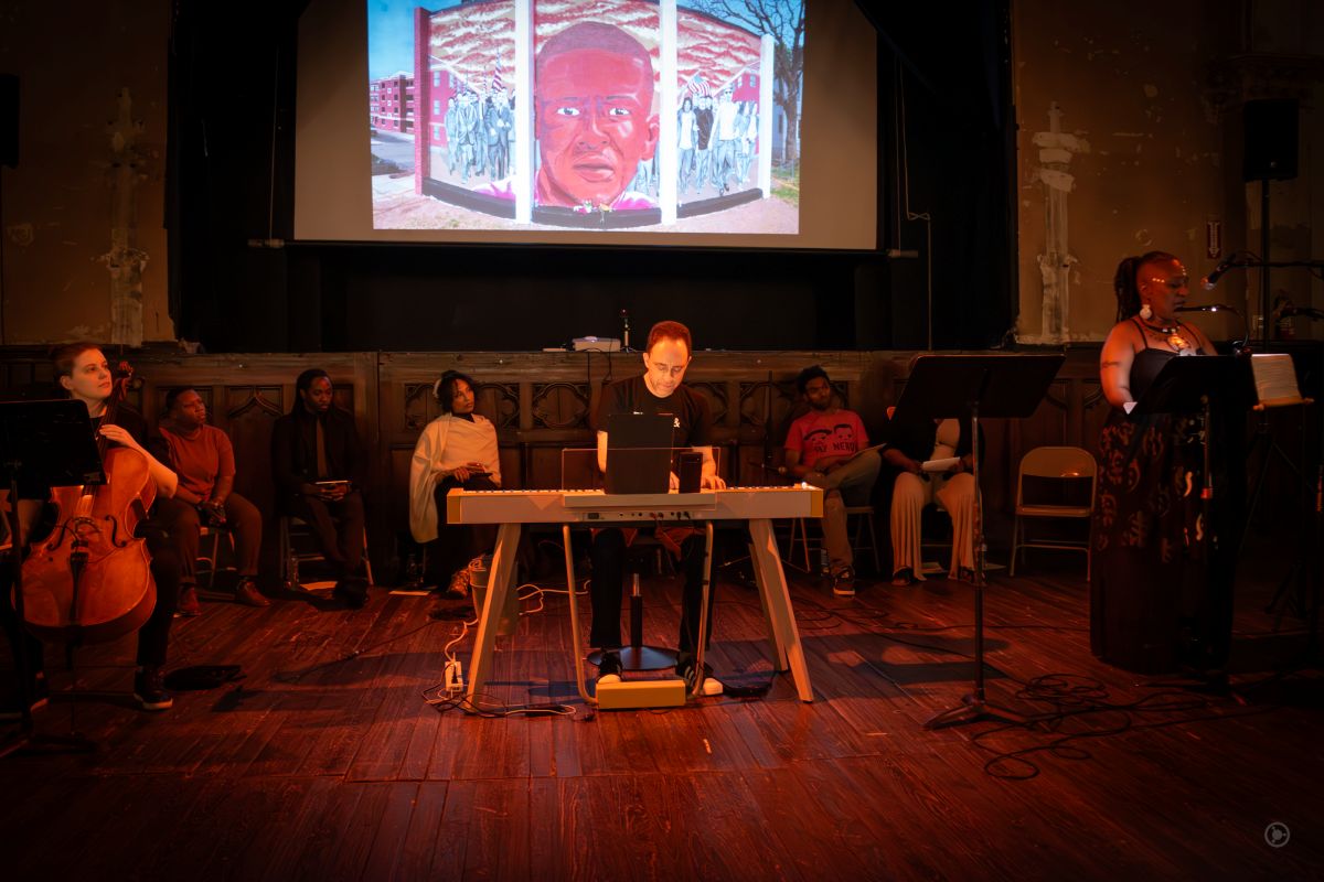 A stage performance features cellist Lavena Johanson on the left, pianist and composer Judah Adashi at center, and activist Erricka Bridgeford standing at a microphone on the right. They perform in a dimly lit venue with a projected image of a mural of Freddie Gray displayed behind them. Several seated participants are in the background, suggesting a collaborative, multimedia tribute or performance centered on social justice.