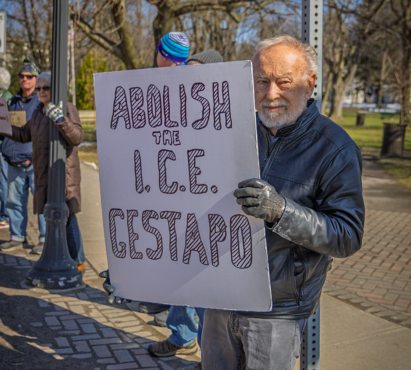 An older man with a white beard and gray hair stands on a brick sidewalk holding a large hand-lettered sign that reads "ABOLISH the I.C.E. GESTAPO." He wears a black leather jacket, gray leather gloves, and gray jeans. Behind him, other protesters stand near a lamppost and metal signpost, bundled in winter coats and knit hats. Bare trees and a clear blue sky fill the background, with long afternoon shadows stretching across the pavement.